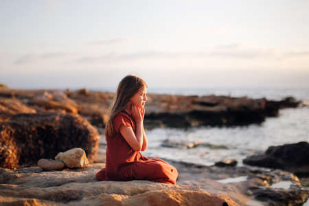A European teenage girl in a dress sits on stones on the seashore, peace and quiet, a lonely child looks at the seaの写真素材