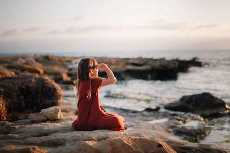 A European teenage girl in a dress sits on stones on the seashore, peace and quiet, a lonely child looks at the seaの写真素材
