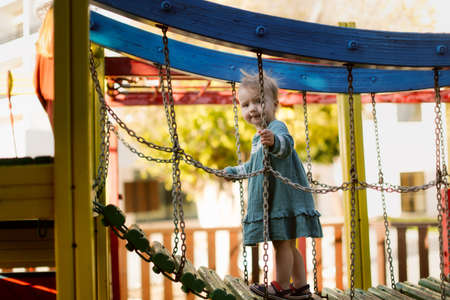 Child girl in a casual dress on the playground climbs up the hinged ladder, summer playgroundの写真素材
