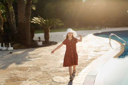 A child girl teenager in a casual dress with a hat walks near the pool in the evening at sunset.の写真素材