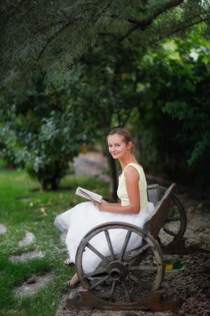 Cute european girl woman with a book on a garden bench under a willow tree, a beautiful garden and a wooden bench made of wagon wheels, landscape art and a beautiful well-groomed gardenの写真素材