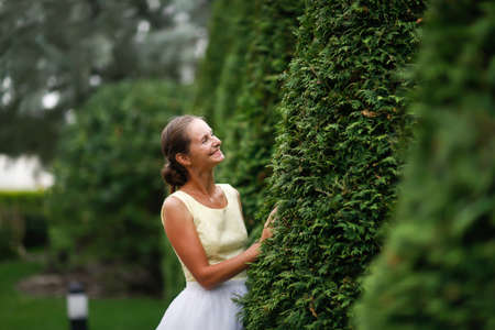 Beautiful young woman in a white tulle skirt near a pyramidal thuja, garden topiary art. Pretty european woman gardener in a festive dressの写真素材