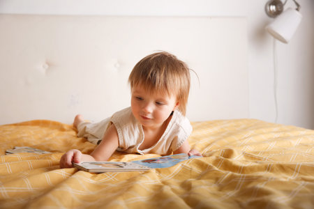 kid flips through a book on bed in bedroom, girl Toddler in overalls lies on bed with book