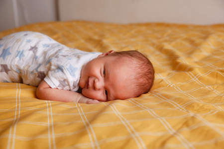 Cute bald chubby baby 2 months old lies on his stomach on yellow sheet on bed in bedroomの写真素材