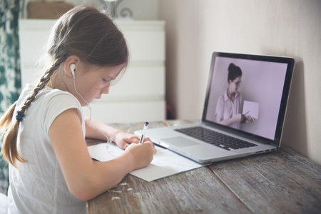 Cute european kid girl with two pigtails studying at home at the table with a laptop and headphones,...