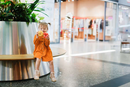 Toddler girl eats donut in supermarket on bench, snack with pastries shopping. Cute child girl in long sleeve mustard dress plays with yellow and pink donutsの写真素材