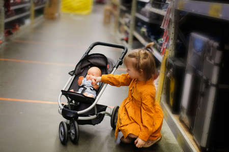 Cute child girl in mustard dress with doll in puppet stroller in store, kid is walking and playing in hardware store, play and care about dollの写真素材