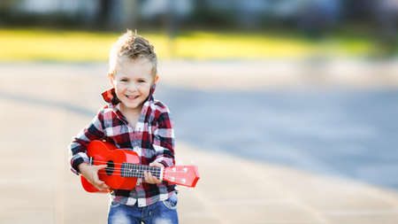Stylish european boy in a shirt plays the red ukulele on the asphalt in the city. Fashionable blond child on a walk. musical childの写真素材