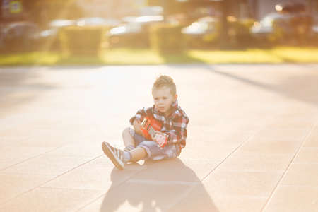 Stylish european boy in a shirt plays the red ukulele on the asphalt in the city. Fashionable blond child on a walk. musical childの写真素材