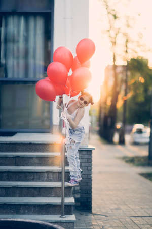 European child preteen girl in torn jeans with red balloons on stairs sunny summer day. Child in sunglasses at sunset on city streets, generation Z, happy birthdayの写真素材