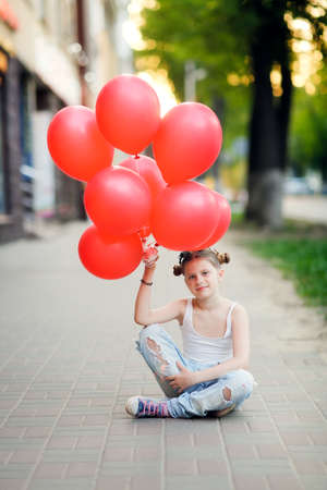 Girl child preteen in jeans and a white t-shirt sits on the sidewalk with a bunch of red balloons, generation Z, lonely holidayの写真素材