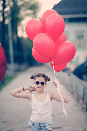 stylish European preteen girl with red balloons in torn jeans on sidewalk in city on summer day child in sunglasses at sunset on streets of town.の写真素材