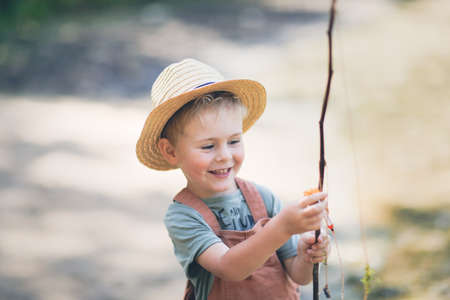 Preschooler boy plays fishing, child fishes with stick near lake in countryside, games in nature and childhood without gadgets, summer holidaysの写真素材