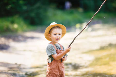 Preschooler boy in yellow rubber boots plays fishing, child fishes with stick near lake in countryside, games in nature and childhood without gadgets, summer holidaysの写真素材