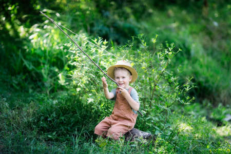 cute european boy in hat and overalls in summer with stick fishing rod fishes in the grass near the lake, plays fishing. happy childhood in countrysideの写真素材