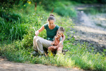Mom and son in park among grass are playing fishing with stick and fishing rod. Mom hugs boy, helps him. concept of happy childhood in nature and happy single parentの写真素材