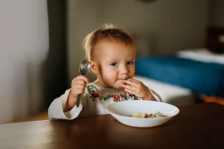 Cute toddler toddler eats porridge for breakfast at the table in the real interior, child with a spoon and plate on a dark background, lifestyleの写真素材