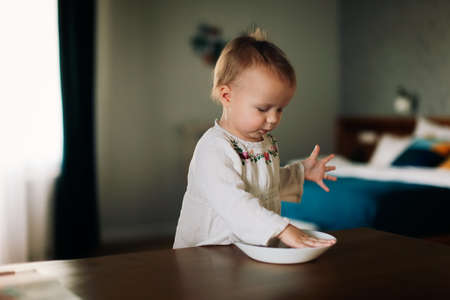 Cute baby kid Toddler eats porridge for breakfast at the table in the real interior, a little child eats hands from a plate on a dark background, lifestyleの写真素材