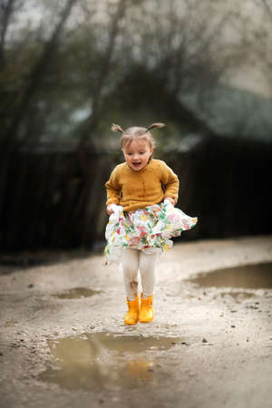 Cute funny girl child toddler jumping through the puddles in yellow sweater and rubber boots, happy...