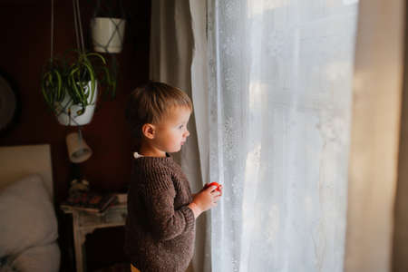 Cute kid toddler in warm sweater near the window in cozy interior, bedroom in warm colorsの写真素材
