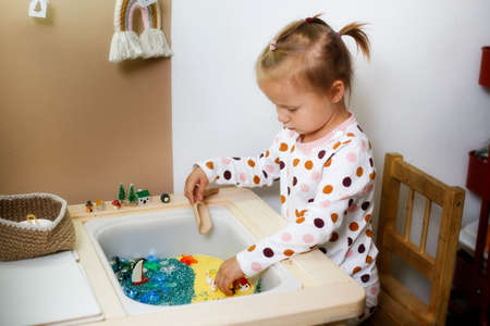 Cute little toddler girl in pajamas plays with box for sensory development, sea theme with sand and stones, development of fine motor skills in children, children's toys in sensory tableの写真素材