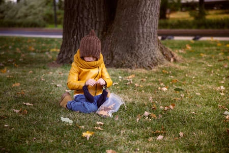 A little girl in a yellow jacket and a knitted hat sits under a tree in an autumn park.の写真素材