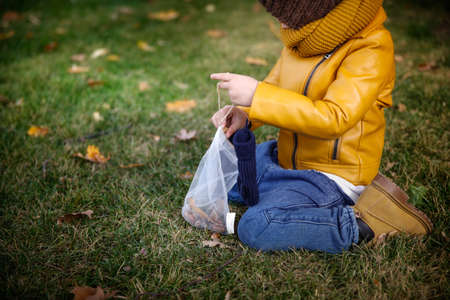 A girl in a yellow jacket and blue jeans sits on the grass in an autumn park and holds a bag with clothes.の写真素材