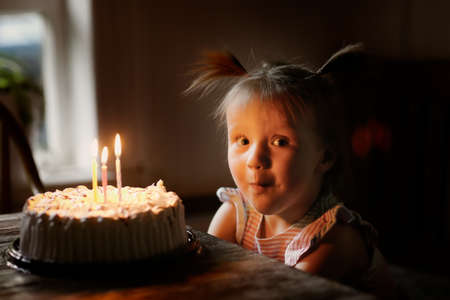 Funny baby toddler girl with two ponytails blows out three candles on the cake, dark style. Children's birthday conceptの写真素材