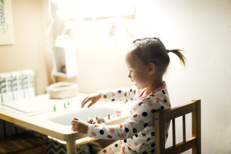 Funny little toddler girl child with a ponytail in pajamas plays at the table in the nursery, independent games and earlier development of childrenの写真素材