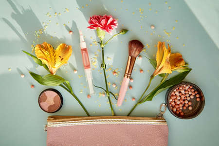 Creative feminine clutch with cosmetic products, accessories, brushes for makeup and powder. High angle hard shadows. Beauty concept. Top view, copy space blue background. Surreal flowers.の写真素材