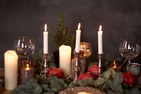 Elegant Christmas table setting with cozy candles, plates, pomegranate and branch of fir tree close...