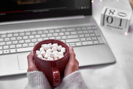 Cozy home office: laptop and red cup with marshmallows close up. Festive working atmosphere with garlands.1st of December. Winter cozy home atmosphere. Top view.の写真素材
