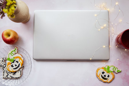 Cozy home office in Halloween: laptop and red cup, homemade cookies in shape of cute pumpkins. Atmospheric working atmosphere with garlands. Copy space.の写真素材