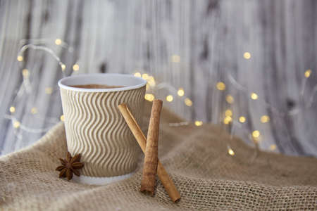 Wooden and sackcloth organic festive Christmas background design with copy space. Cardboard cup with hot beverage, cinnamon sticks and anise star. Garlands bokeh in the backgroundの写真素材