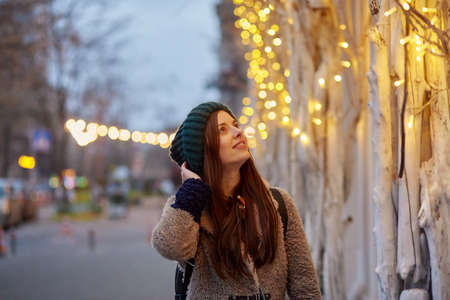 Dreamy caucasian woman walking in the street. City lights. Winter casual clothes. Festive city at...