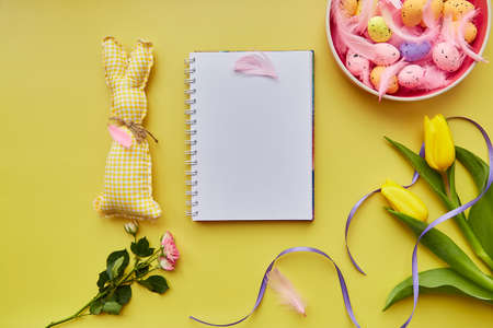 Festive Easter mock up notepad. Pink bowl with colorful eggs, pink feathers, handmade cute bunny and flowers. Happy Easter concept. Post card mockup on yellow backgroundの写真素材