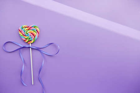 Rainbow heart shaped sweet lollipop on trendy violet background with festive ribbon and shadows....