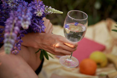 Woman holds a wine glass with purple lupinus at sunset. summer picnic. Wellness, natural concept. Adaptogenic ayuverdic drink concept. Conscious consumption. Extra wide banner. purple place for textの写真素材