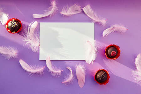 White mock up on purple background with shadows: chocolate sweets and pink feathers. Anniversary, happy birthday, wedding, mothers day wishing. Holiday present concept. Purple pattern, copy space.の写真素材