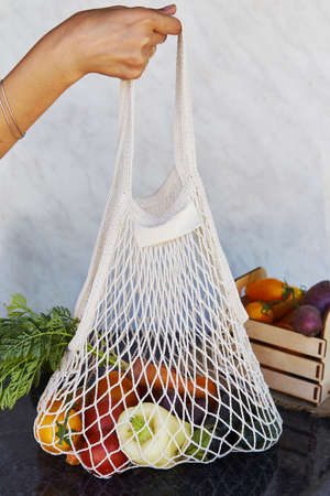 Woman holds eco shopping reusable bag full of fresh vegetables - tomatoes, purple potatoes,...