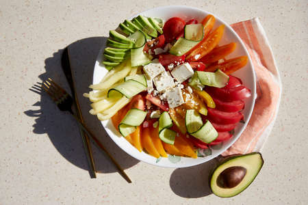 Healthy seasonal salad with red, orange and yellow tomatoes, sweet pepper, avocado, cucumbers, feta cheese, sesame under hard shadows. Natural, organic food, proper nutrition.の写真素材