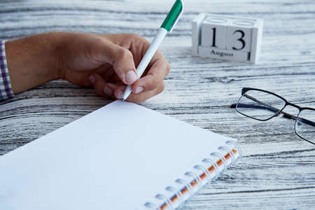 Man uses his left hand to wright in a notebook close up. Glasess and calendar with 13th of August - Day of Handlefters. Conceptual photo of lefties day. Mock up notepad. Copy space.の写真素材