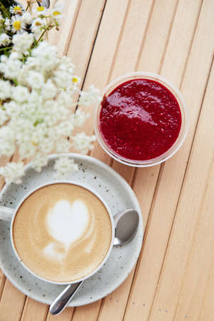 Cappuccino in a white ceramic cup with heart and raspberry jam dessert in a disposable cup - a...