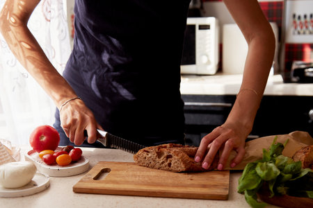 Woman is cutting a French fresh crispy baguette in the kitchen under shadows. Basil, tomatoes and...