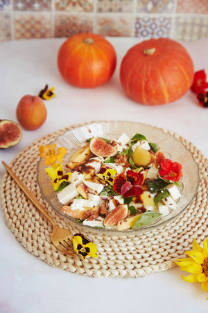 Fresh autumn vegetarian salad with edible flowers, figs, peach, greens and feta on ceramic tile background with fall pumpkin decorations.Selective focusの写真素材