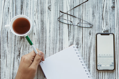 Man writes in a notebook with his left hand in a notepad, phone screen mockup. Glass and cup of tea. Conceptual photo of Happy left hander's day.の写真素材
