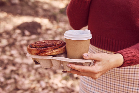 Woman walking in the park with cinnabon bun and coffee. Aesthetic autumn breakfast.の写真素材