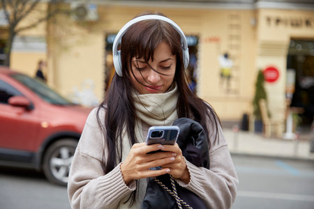 Happy concentrated young woman in earphones using smartphone outdoor. Freelance, influencer, chatting concept.の写真素材