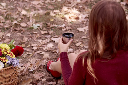 Woman hand holds a tea cup. Aesthetic autumn picnic ideas in the park among flowers. Cozy autumn breakfast.の写真素材