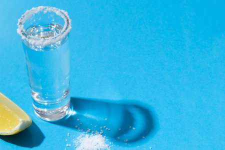 Single shooter glass of tequila on blue background with salt and lemon. Concept party. Close up copy...